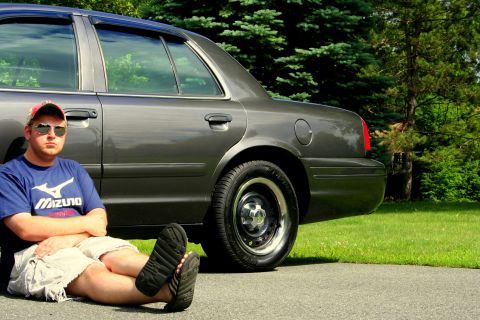 2000 Ford Crown Victoria - Man sits beside a 2000 Ford Crown Victoria parked on a paved surface with grass and trees in the background.