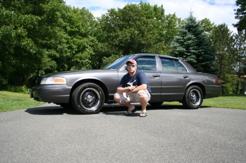 2000 Ford Crown Victoria - A man poses in front of a gray sedan on a paved driveway with trees in the background.