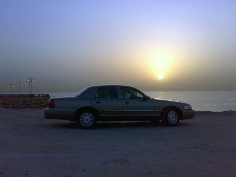 2007 Mercury Grand Marquis - A sedan is parked on a sandy area near the water as the sun sets on the horizon.