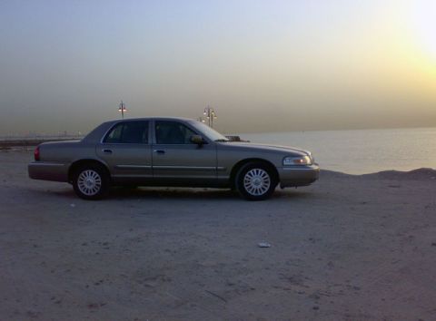 2007 Mercury Grand Marquis - A tan sedan sits on a sandy shore as the sun sets over the water.