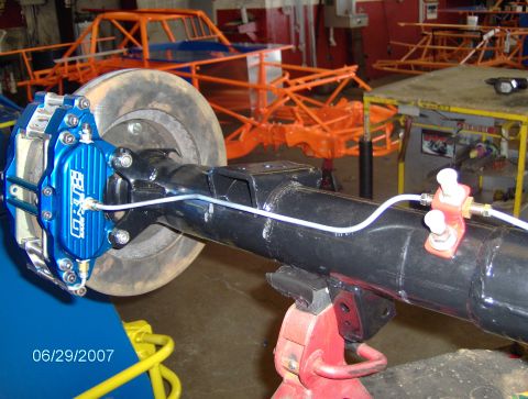 1978 Ford Bronco - Close-up of a rear axle assembly a blue brake caliper with "GRUDGE" branding. Multiple unfinished race car chassis are visible in the backg…