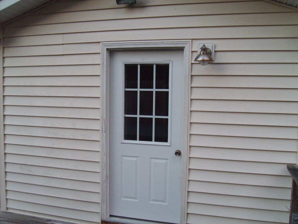 White door with a nine-pane window, mounted on a beige vinyl-sided wall with a light fixture above