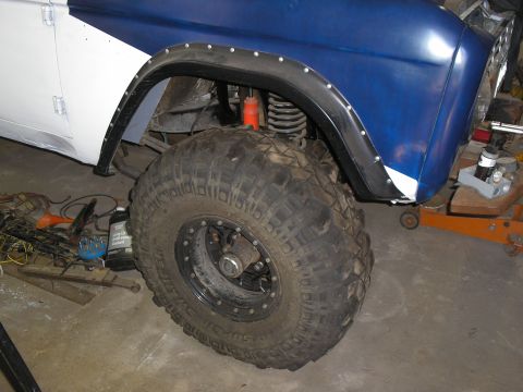 1966 Ford Bronco - Passenger-side front wheel well of a blue and white early Bronco in a garage, showing a black riveted fender flare, coil spring with orange…