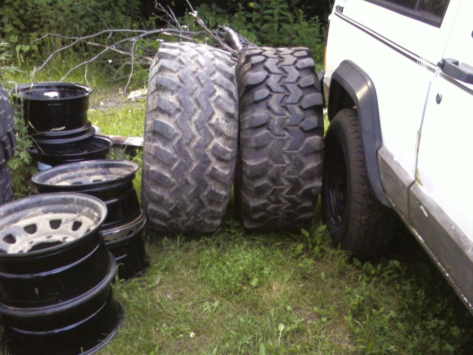 Pile of off-road tires and rims next to a white vehicle