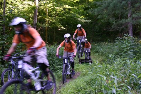 1997 Ford Probe - Four mountain bikers in orange shirts and helmets ride down a dirt path surrounded by trees and greenery.