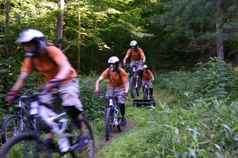 Group of people riding mountain bikes on a wooded trail, some going over a small ramp