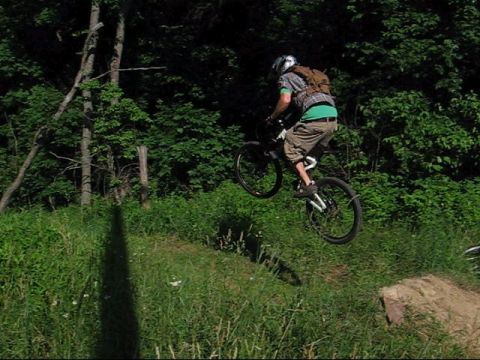 1997 Ford Probe - A mountain biker performs a jump in a wooded area, with a shadow cast on the grassy slope below.