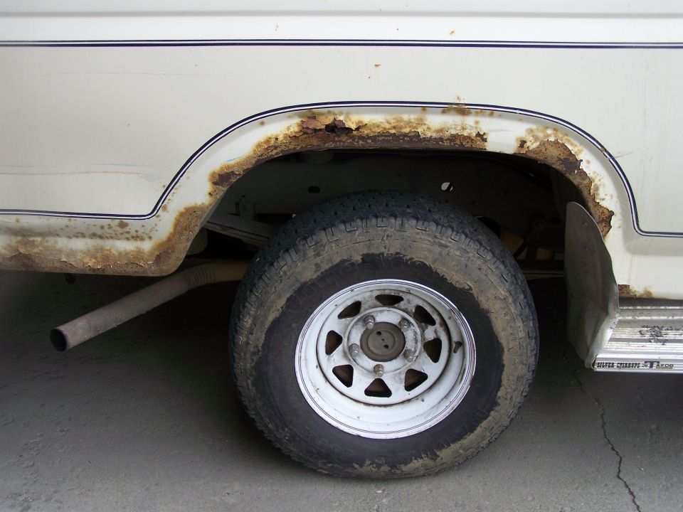Rust damage around rear wheel arch of white Bronco with steel spoke wheel and hanging exhaust pipe