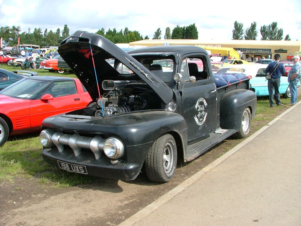 Black 1950s pickup truck with exposed engine, custom grille, and license plate 198 UXS