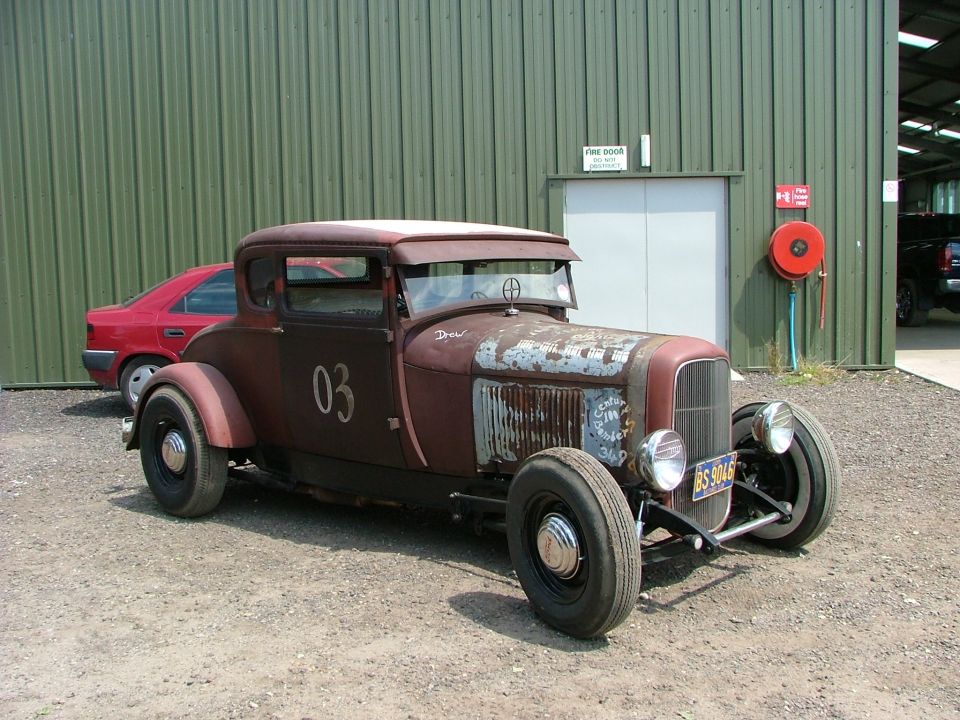 Rusty hot rod coupe with number 03 on the door, custom grille, and exposed front suspension. A red sedan is parked behind it
