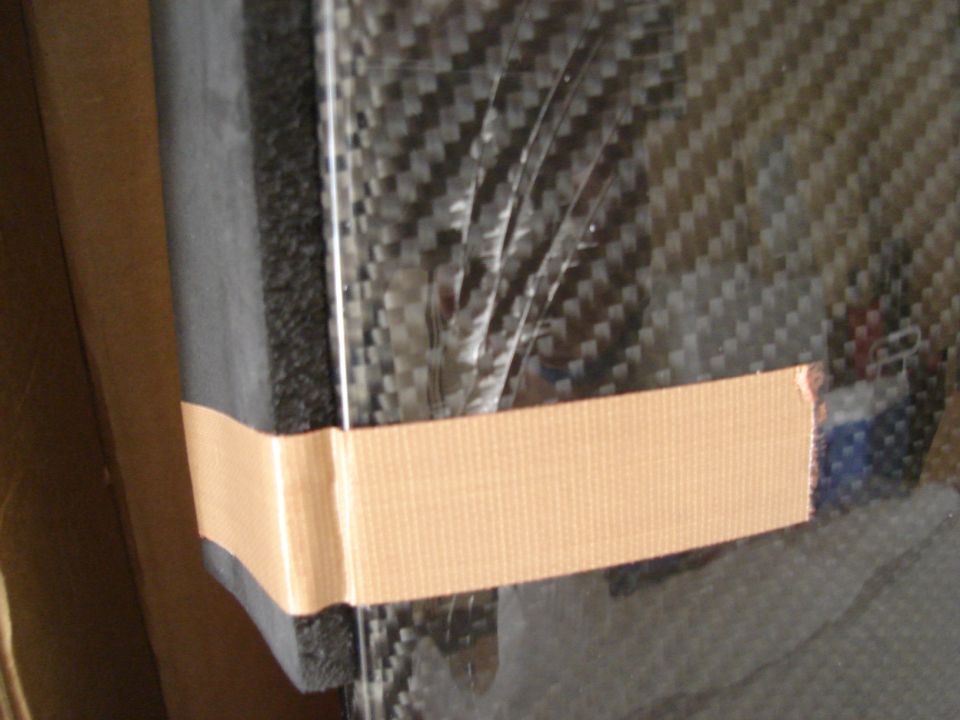 Carbon fiber panel secured with tan tape, showing a woven texture and plastic film