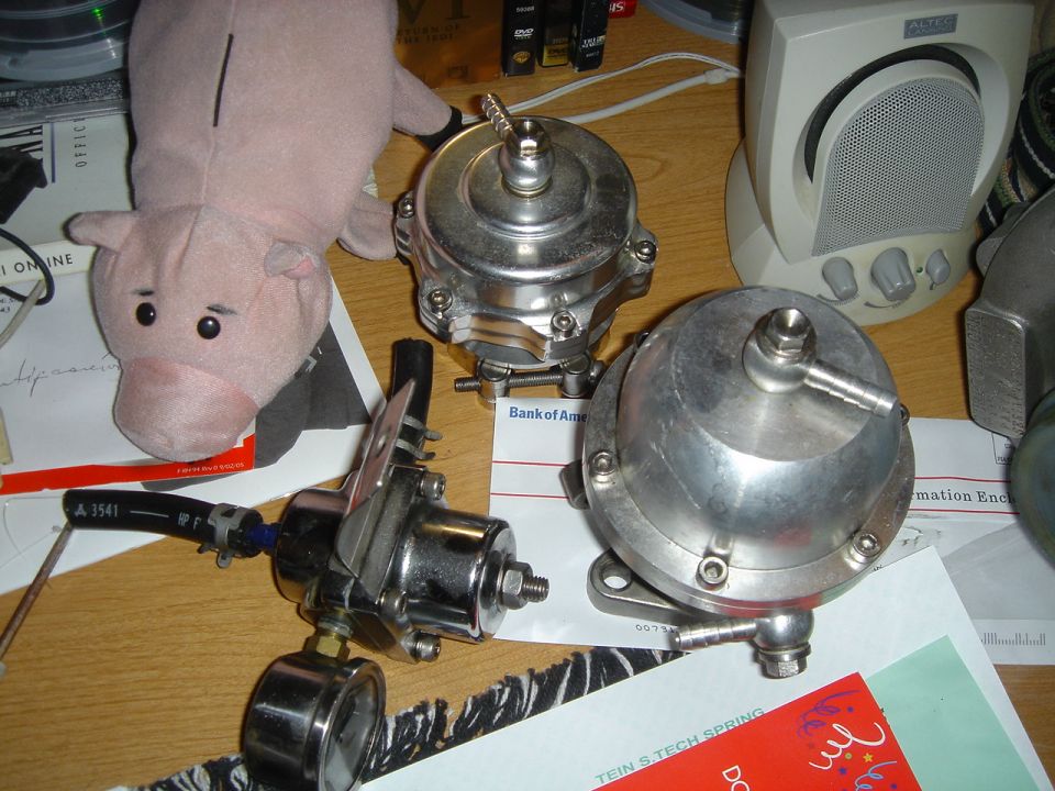 three shiny metal automotive parts, one with a gauge, and a pink stuffed pig on a desk with papers