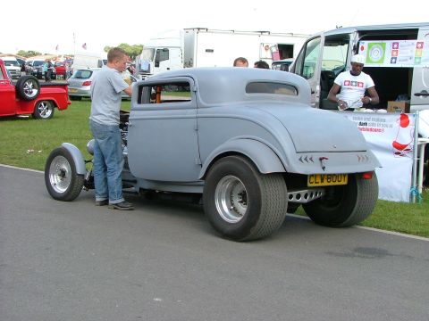 2001 Ford Fiesta - A custom hot rod coupe is displayed outdoors with a man attending to it near a vendor booth.