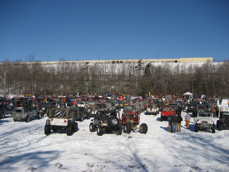 many off-road vehicles parked in a snowy lot with trees and a building in the background