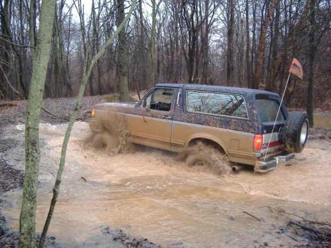 1991 Ford Bronco - This Ford Bronco is navigating a challenging muddy terrain, kicking up water as it goes.