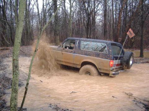 1991 Ford Bronco - A Ford Bronco navigates a muddy off-road trail, kicking up water and dirt.