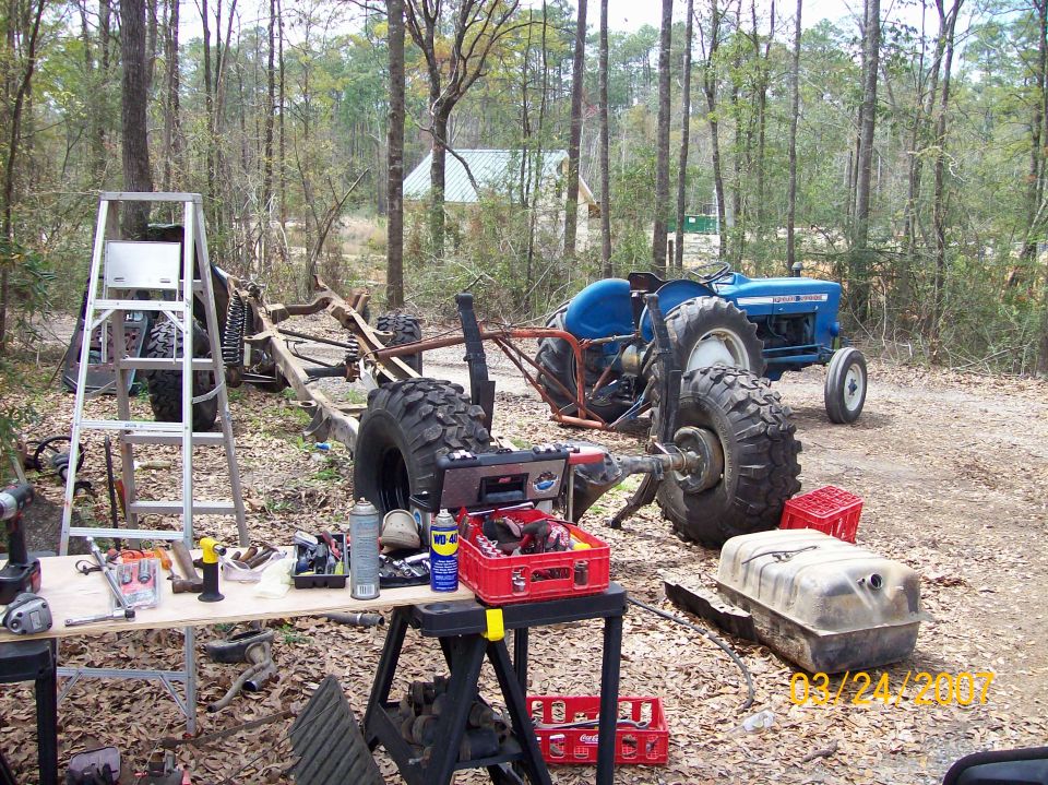 Tractor chassis with large tires, tools, and a blue tractor in the background