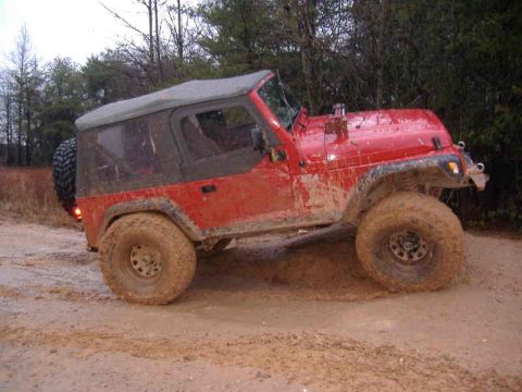 1991 Ford Bronco - Muddy red Jeep Wrangler with a soft top is stuck in a muddy trail.