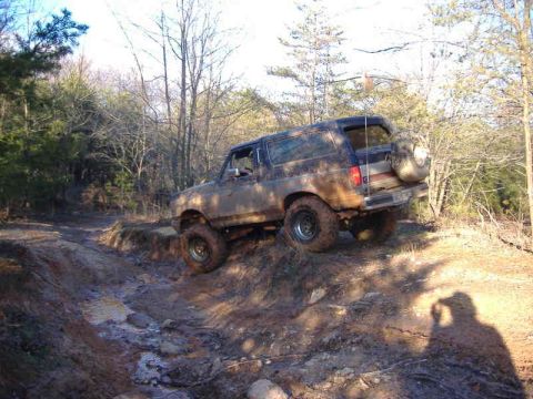 1991 Ford Bronco - A dark, mud-covered SUV navigates a challenging off-road trail with muddy puddles and steep inclines.