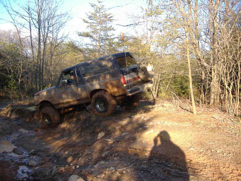Muddy 1991 Ford Bronco climbing a dirt hill with trees in the background
