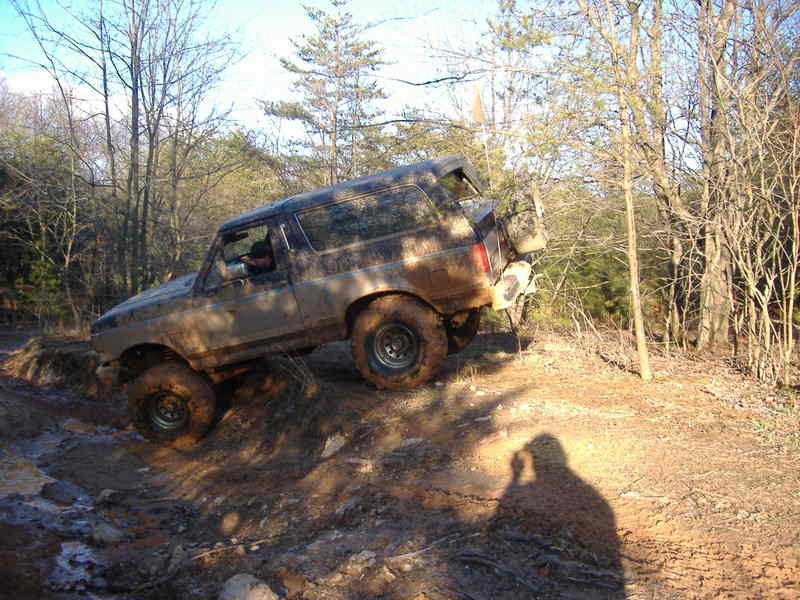 Muddy 1991 Ford Bronco climbing a dirt hill in a wooded area. The spare tire is mounted on the rear