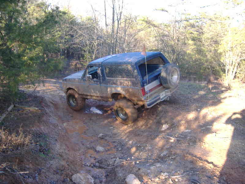 Muddy 1991 Ford Bronco driving on a dirt trail with trees around