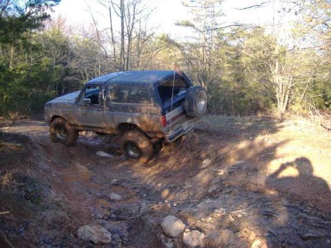 1991 Ford Bronco - A muddy Ford Bronco navigates a challenging off-road trail with trees in the background.