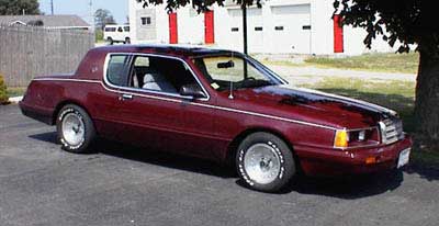 1986 Mercury Cougar - A maroon 1986 Mercury Cougar LS coupe is parked on asphalt in front of a building with a tree to its right.