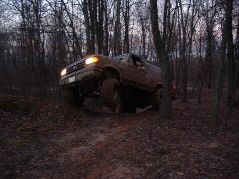 1991 Ford Bronco - A brown Ford Bronco navigates a muddy, wooded trail with one wheel lifted off the ground.