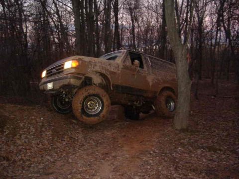 1991 Ford Bronco - A heavily muddied SUV navigates a wooded trail during daylight hours.