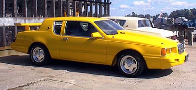 1986 Mercury Cougar - A yellow coupe with a prominent rear spoiler is parked outdoors next to another vehicle.
