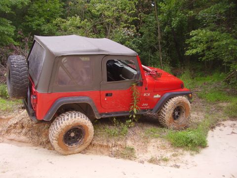 1991 Ford Bronco - A red Jeep Wrangler navigates a muddy off-road trail with its soft top deployed.