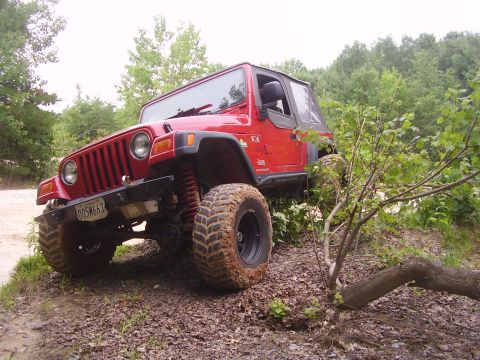 1991 Ford Bronco - A red Jeep Wrangler navigates a challenging off-road course with muddy tires and dense foliage.