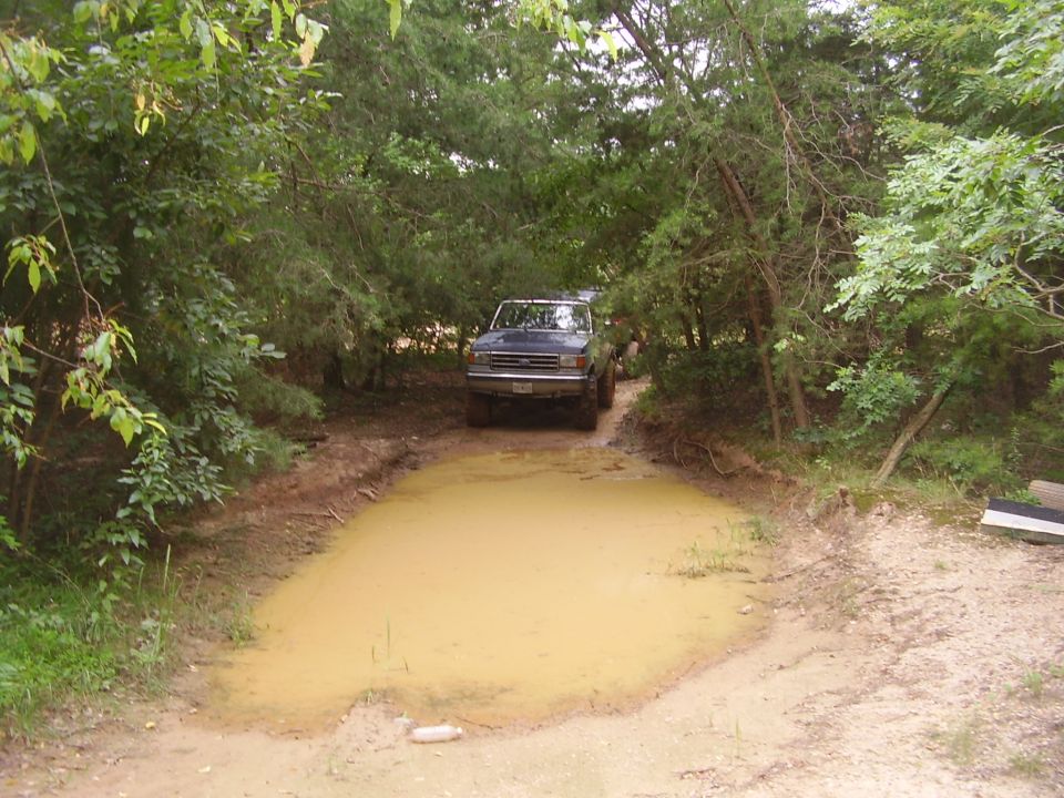Dark blue Ford Bronco approaching a large mud puddle on a dirt path surrounded by trees