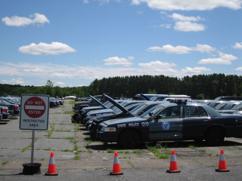 1991 Ford LTD Crown Victoria - Parked police vehicles in a restricted area lot under a cloudy blue sky.