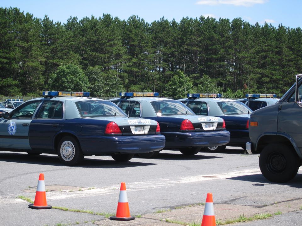 Several police cars parked in a lot with trees in the background. One van is on the right