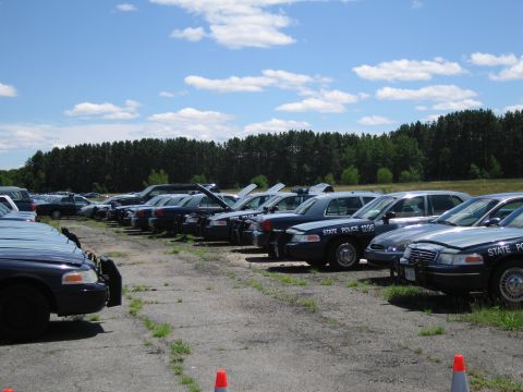 1991 Ford LTD Crown Victoria - A line of dark blue police cruisers are parked in an outdoor lot under a partly cloudy sky.