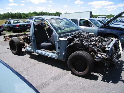 1991 Ford LTD Crown Victoria - A heavily damaged car chassis sits in a junkyard with other vehicles in the background.