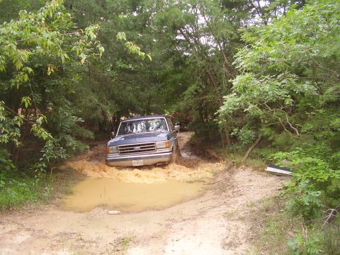 1991 Ford Bronco - A blue Ford F-Series truck navigates a muddy obstacle on a wooded trail.
