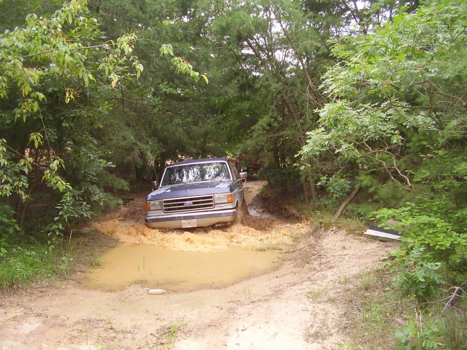 Blue Ford F-Series truck driving through a muddy puddle on a dirt road