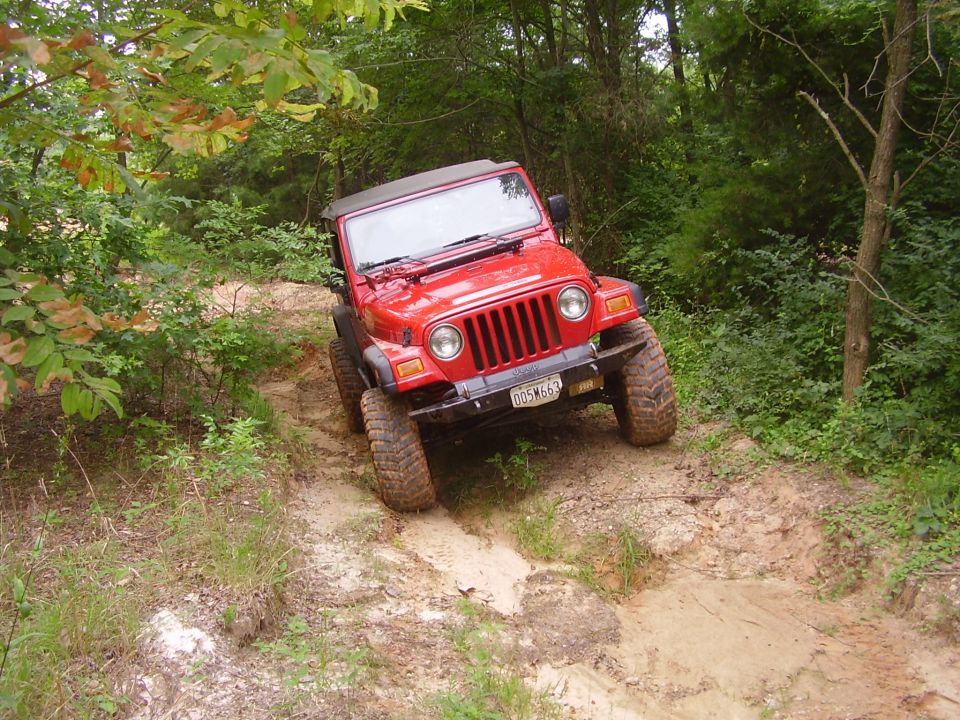 Red Jeep Wrangler TJ model driving off-road on a sandy trail with trees on either side. License plate reads D05M663