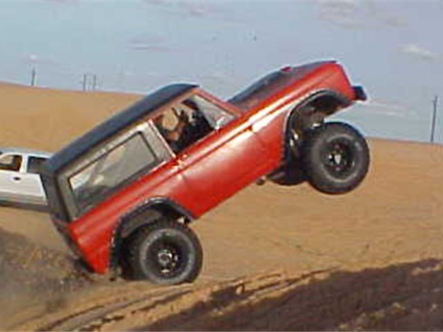 Red SUV airborne over a sandy dune with a white vehicle behind it
