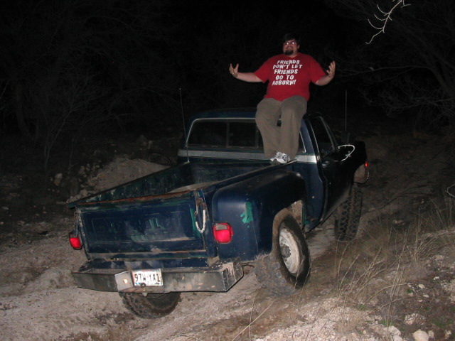 Man in red t-shirt stands on the tailgate of a blue pickup truck, off-road at night. License plate reads 97-111