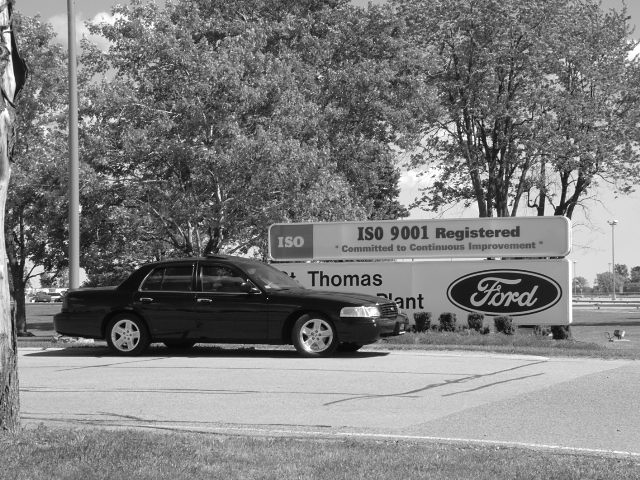 Black sedan parked in profile in front of a Ford plant sign with trees behind.