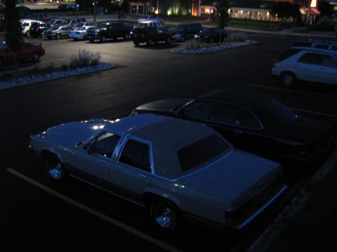 1988 Mercury Grand Marquis - Elevated night shot looking down on a light-colored boxy four-door sedan with a vinyl half-roof and chrome slotted wheels, parked in a lot…