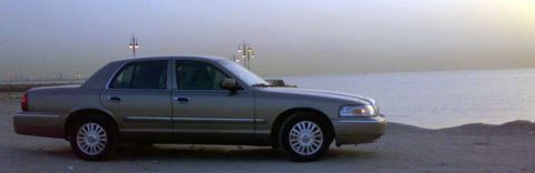 2007 Mercury Grand Marquis - A silver-gold Mercury Grand Marquis full-size sedan photographed in profile on a sandy waterfront lot at dusk, with chrome-look multi-spoke…