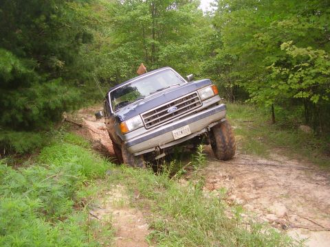 1991 Ford Bronco - This Ford truck is navigating a muddy, off-road path surrounded by dense foliage.