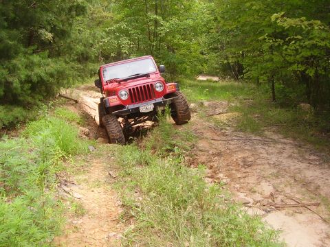1991 Ford Bronco - A red Jeep Wrangler tackles a challenging off-road course, its large tires digging into the dirt and mud.