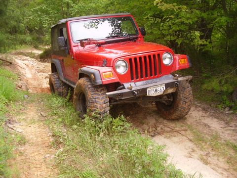 1991 Ford Bronco - A red Jeep Wrangler navigates a muddy, wooded trail with its tires kicking up dirt.
