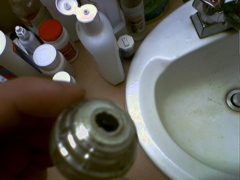 2001 Ford Crown Victoria - A close-up view of a faucet handle part is held in front of a bathroom sink, surrounded by various bottles and containers.